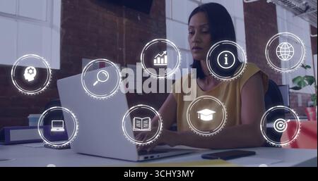 Donna matura che indossa la mostarda sulla scrivania dell'ufficio, sul laptop e sulle icone digitali sovrappongono lo spazio di copia Foto Stock