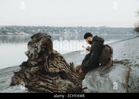 Giovane con zaino seduto sulla riva del Danubio a Novi Sad, mentre usa il telefono durante una fredda giornata invernale. Foto Stock