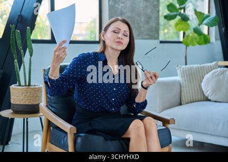 Donna matura che soffre di sintomi di febbre menopausa, agitando le carte Foto Stock