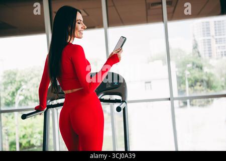 Giovane donna in un abbigliamento sportivo rosso brillante che utilizza uno smartphone mentre si esercita su un tapis roulant in una palestra moderna e luminosa Foto Stock