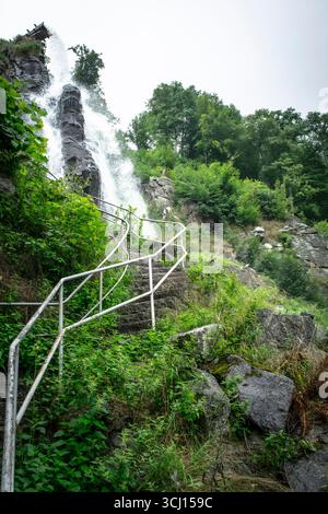 Spettacolare cascata a cascata accanto a scale in pietra con ringhiere rustiche, circondata da vegetazione lussureggiante e nebbia, evocando avventura e meraviglia naturale. Foto Stock