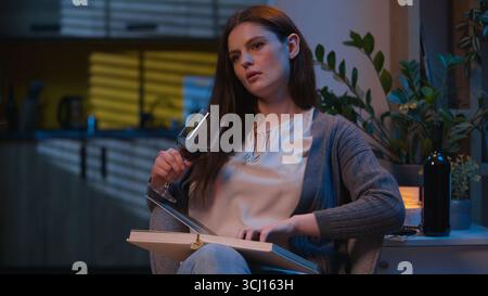 Donna triste e premurosa ragazza sola notte a casa guardare album fotografico bere vino di bicchiere rilassarsi in appartamento serale immagini ricordi pensando sconvolta Foto Stock