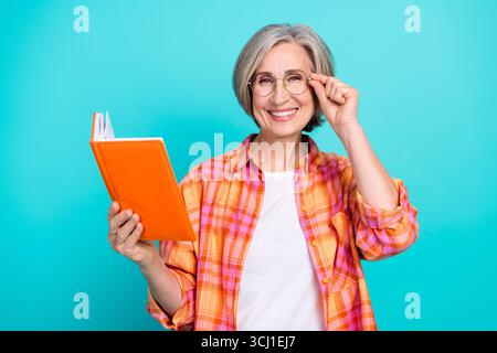 Donna matura sorridente che tiene in mano occhiali da regolazione su un vivace sfondo color tè che mette in mostra relax e conoscenza Foto Stock