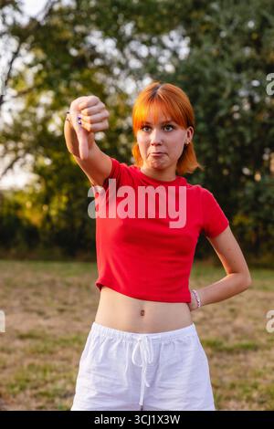 Una giovane donna che mostra un gesto con i pollici giù nel parco che esprime disapprovazione Foto Stock