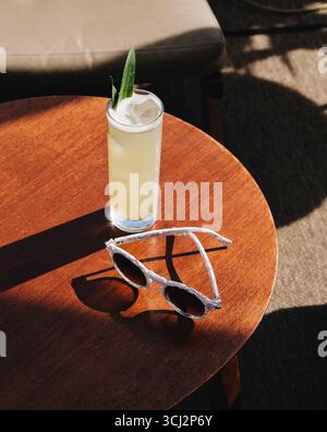 classico scenario estivo di vacanza caratterizzato da un alto e rinfrescante cocktail tropicale e un elegante paio di occhiali da sole appoggiati su un tavolo di legno illuminato dal sole. Foto Stock
