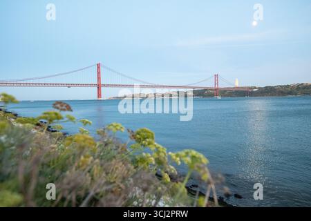 Una splendida foto panoramica del fiume Tago, con l'iconico ponte rosso 25 de Abril e il lontano monumento Cristo Rei. La fotografia cattura Foto Stock