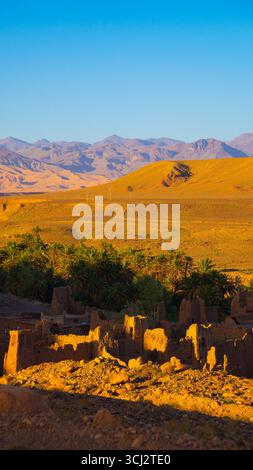 Tradizionale villaggio marocchino con palme e case in mattoni adagiate contro la spettacolare catena montuosa dell'Atlante sotto il cielo sereno Foto Stock