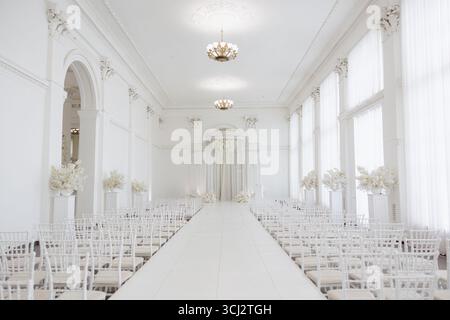 Un elegante spazio per cerimonie nuziali pieno di decorazioni floreali bianche. File di sedie si affacciano su un corridoio luminoso sotto splendidi lampadari. Foto Stock