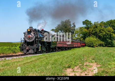 Strasburg, Pennsylvania, 8-9-2025 - Una locomotiva a vapore d'epoca trasporta un lungo treno passeggeri attraverso campi verdi sotto un cielo azzurro. Il motore emette fumo nero, creando un'atmosfera nostalgica. Foto Stock