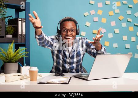 Imprenditore nero che indossa le cuffie, tira con le braccia alzate e sorride ampio, esprimendo entusiasmo dopo aver concluso un accordo commerciale. Il manager ha festeggiato con gioia l'approvazione del progetto di avvio. Foto Stock