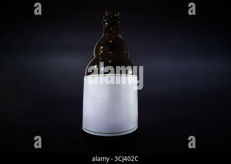 Una bottiglia di birra marrone scuro con etichetta bianca semplice è isolata su uno sfondo nero dello studio, offrendo uno spazio di copia pulito per il branding personalizzato Foto Stock