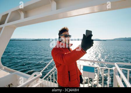 Turista con gli occhiali da sole che fa selfie davanti al paesaggio dell'oceano tenendo il cellulare in piedi sulla barca. Bearded Guy che sta videochiamando usando il cellulare Foto Stock