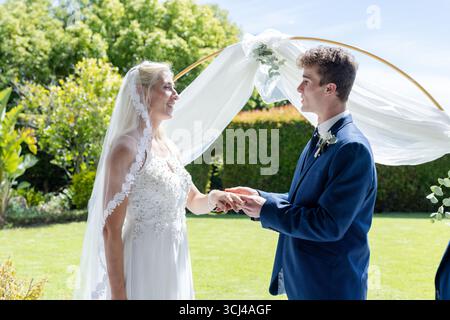 Sposa e sposo che si scambiano le promesse all'aperto sotto un elegante arco floreale, sorridendo calorosamente Foto Stock