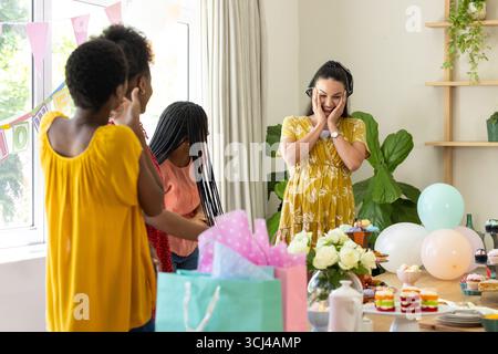 Donna sorprendente con torta di compleanno e regali, diverse amiche che festeggiano a casa Foto Stock