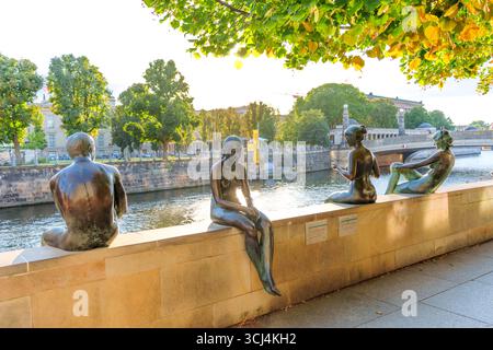 Berlino, Germania - 17 agosto 2025: Gruppo di statue in bronzo raffiguranti tre ragazze e un ragazzo seduti lungo il fiume Sprea, circondati da una vegetazione lussureggiante Foto Stock