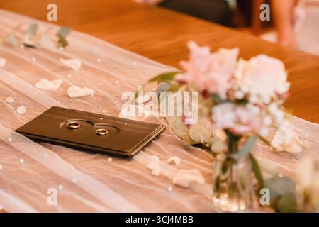 Le fedi nuziali sono elegantemente esposte su un vassoio scuro con petali floreali sparsi e delicati dettagli in tessuto. I fiori rosa tenui esaltano il romantico Foto Stock