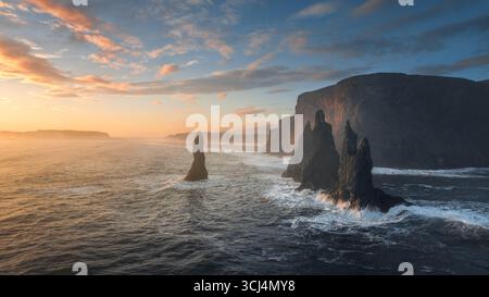 Veduta aerea delle scogliere frastagliate incontra il turbolento oceano sotto un cielo che si illumina con le sfumature ardenti del tramonto, Reynisfjara, Islanda. Foto Stock