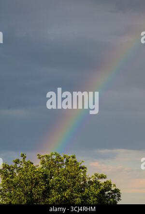 Dopo la pioggia, un vivido doppio arcobaleno appare sopra un villaggio illuminato dal sole, contrastando splendidamente con il cielo grigio mostruoso. Foto Stock