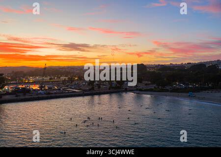 Veduta aerea del tranquillo porto che rispecchia il tramonto ardente, dove l'oceano incontra la riva sotto un cielo che si illumina di tonalità arancio e rosa, Dana Point, California, Stati Uniti. Foto Stock