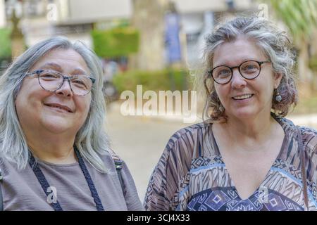 Due donne mature con capelli e occhiali grigi sorridono all'aperto, magari trascorrendo una piacevole giornata insieme Foto Stock
