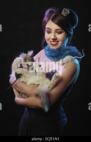 La ragazza felice con il trucco della Regina del mouse abbraccia il gatto su sfondo nero, sorridendo Foto Stock
