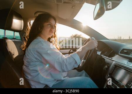 Una giovane donna siede al volante della sua auto durante il tramonto Foto Stock