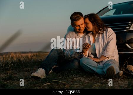 Coppia seduta a terra vicino alla macchina al tramonto Foto Stock