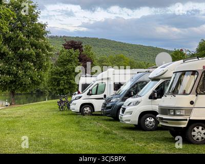 Diversi camper sono parcheggiati in un'area erbosa accanto a un fiume, con una collina verde sullo sfondo, che offre una vista tranquilla e panoramica Foto Stock