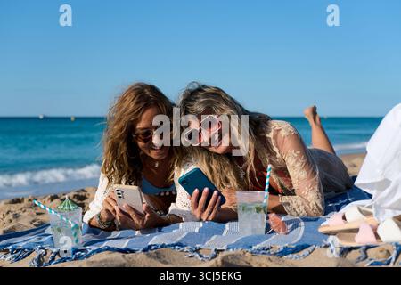 Due donne mature che utilizzano smartphone in spiaggia, si godono le vacanze estive Foto Stock