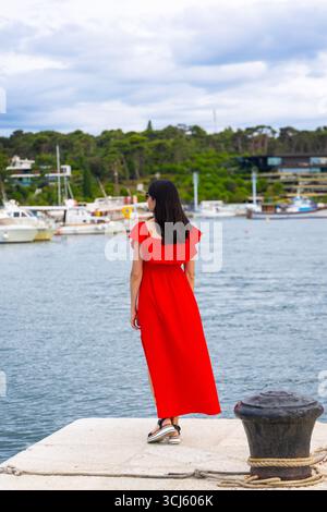 Donna in abito rosso in piedi al molo del porticciolo. Una donna che indossa un lungo abito rosso e sandali si trova in un molo del porto, guardando le barche ormeggiate sotto una Foto Stock
