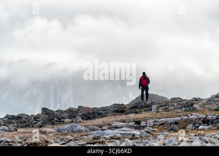 Danes Island Svalbard Person with Rifle Watches for Polar Bears // DANES ISLAND, Svalbard - Una persona si erge su un paesaggio roccioso e arido sull'isola Danes (Danskøya), parte dell'arcipelago norvegese delle Svalbard nell'Oceano Artico. L'isola, con un'area di 40,6 km² (15,7 sq mi), si trova a nord-ovest di Spitsbergen ed è in gran parte disabitata. Negli anni '1630, gli olandesi fondarono una stazione chiamata "Cookery of Harlingen" sul lato nord dell'isola di Danes. Più tardi, nel 1897, la spedizione in mongolfiera artica di S. A. Andrée partì dall'isola di Danes, anche se la spedizione finì tragicamente. Foto Stock