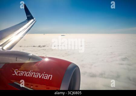 SAS Aircraft Wing over Clouds vicino Longyearbyen Norvegia // NORVEGIA SETTENTRIONALE — l'ala di un aereo SAS Scandinavian Airlines è visibile sopra uno spesso strato di nuvole, con diverse piccole isole che appaiono all'orizzonte. Il cielo azzurro sopra indica un volo ad alta quota. SAS, precedentemente Scandinavian Airlines System, è la compagnia aerea di bandiera di Danimarca, Norvegia e Svezia. Foto Stock