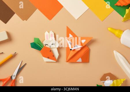Coniglio di carta con figure di origami di carota e volpe sulla scrivania con fogli colorati, forbici, matita, gomma e colla. Giocattolo artigianale per bambini. Foto Stock