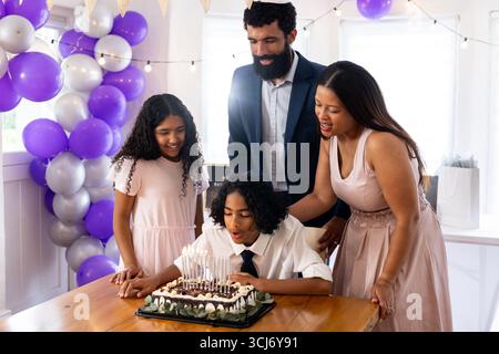 La famiglia che festeggia il compleanno mentre un ragazzo fa saltare le candele sulla torta a casa Foto Stock