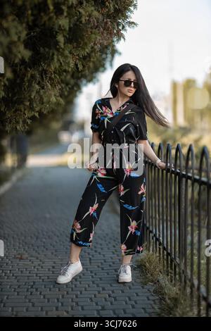 Una giovane donna si trova accanto a un percorso del parco e indossa un elegante outfit floreale nero. Si appoggia contro una recinzione trasudando fiducia nella calda luce del sole circondata dal verde. Foto Stock
