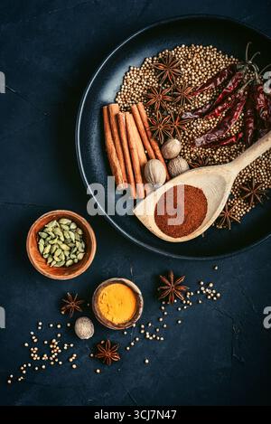 Spezie biologiche miste (coriandolo, noce moscata, stelle di anice, pepe di cayenna, curcuma, paprika, bastoncini di cannella) su piatto di ceramica su sfondo nero, parte superiore Foto Stock