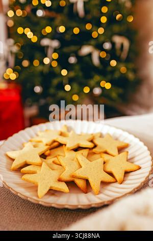 Un piatto di biscotti a forma di stella è esposto di fronte a un albero di Natale splendidamente illuminato, creando un'atmosfera di vacanza calda e festosa. Foto Stock