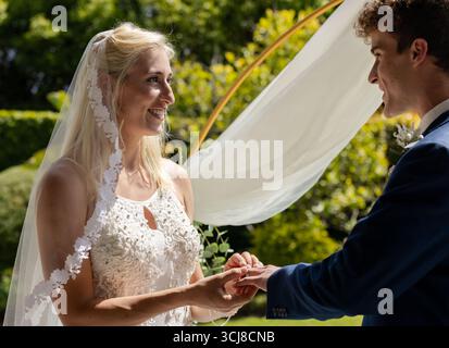 La sposa si scambia anelli con lo sposo all'aperto, entrambi sorridenti alla luce del sole Foto Stock