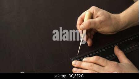 Artigiano che utilizza punteruolo e righello per disegnare linee precise sulla superficie in pelle per la realizzazione di accessori fatti a mano Foto Stock