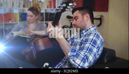 Uomo che suona la chitarra acustica sul divano di casa, con bicicletta, donna che legge libro, copia spazio Foto Stock