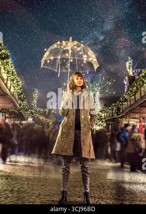 Bella donna alla moda in piedi nel bel mezzo del mercatino di Natale, mentre tiene in mano un ombrello trasparente con luci da favola. Fotografato di notte. Foto Stock