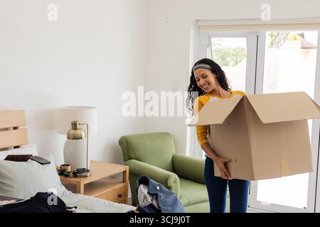 Disimballaggio della scatola di cartone in camera da letto, donna afroamericana che organizza effetti personali, sorridendo Foto Stock