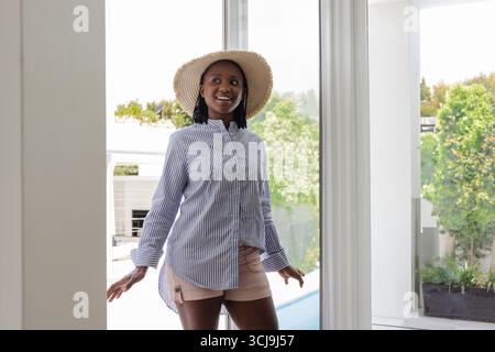 Donna afroamericana che entra attraverso una porta di vetro nell'area della piscina sul retro indossando un cappello da sole in paglia Foto Stock