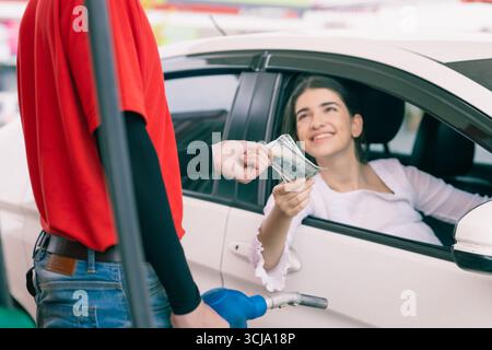 Lavoratore del personale della stazione di servizio che riceve denaro in contanti per il pagamento delle bollette del costo del carburante dal proprietario dell'auto, azienda petrolifera Foto Stock