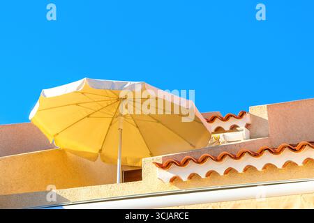 L'ombrello del patio si erge contro il cielo, gettando l'ombra sulle pareti e sulle tegole di terracotta dell'edificio. Bagnato dalla luce del sole, l'ombrellone offre ombra sulla sommità Foto Stock