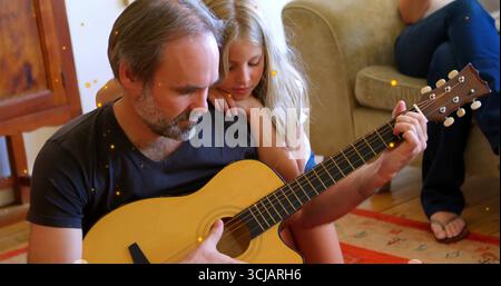 Padre che suona la chitarra acustica in salotto, con figlia appoggiata alla spalla Foto Stock