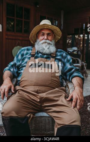 Vecchio agricoltore barbuto con cappello di paglia e tuta che riposano sulla sedia dopo una lunga giornata di lavoro in campagna, godendosi la pace e la tranquillità della sua fattoria Foto Stock