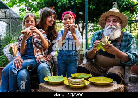 La madre con le figlie e il nonno si godono una sana pausa pranzo insieme, mangiando panini nella loro fattoria biologica, rappresentando i valori della famiglia Foto Stock