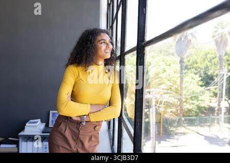 Donna sicura di sé in maglione giallo che guarda fuori dalla finestra dell'ufficio, pianificando obiettivi futuri Foto Stock