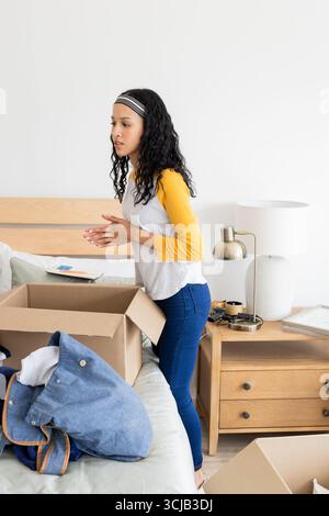 Giovane donna che disimballa le scatole in camera da letto, organizzando gli effetti personali con la massima attenzione Foto Stock